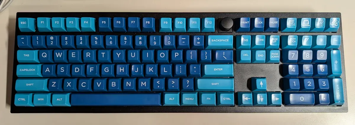 A top-down photo of a mechanical keyboard with gradient blue keys and white lettering, showing all standard keys including…