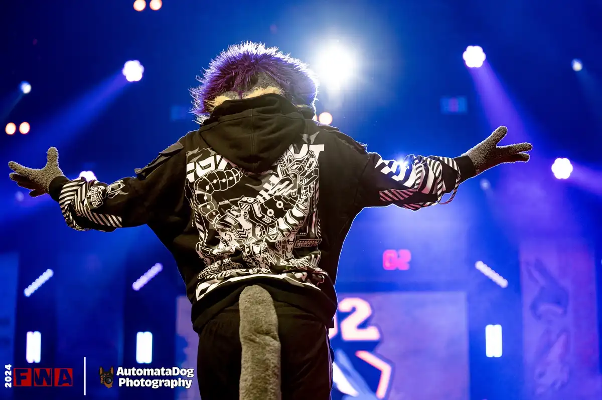 A person with purple spiky hair and a black hoodie with a white graphic design is seen from behind on a stage with blue…