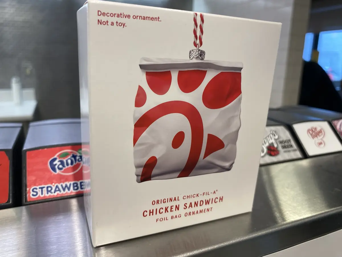 A white box displays a Chick-fil-A chicken sandwich foil bag ornament, featuring the iconic red logo on a white background…