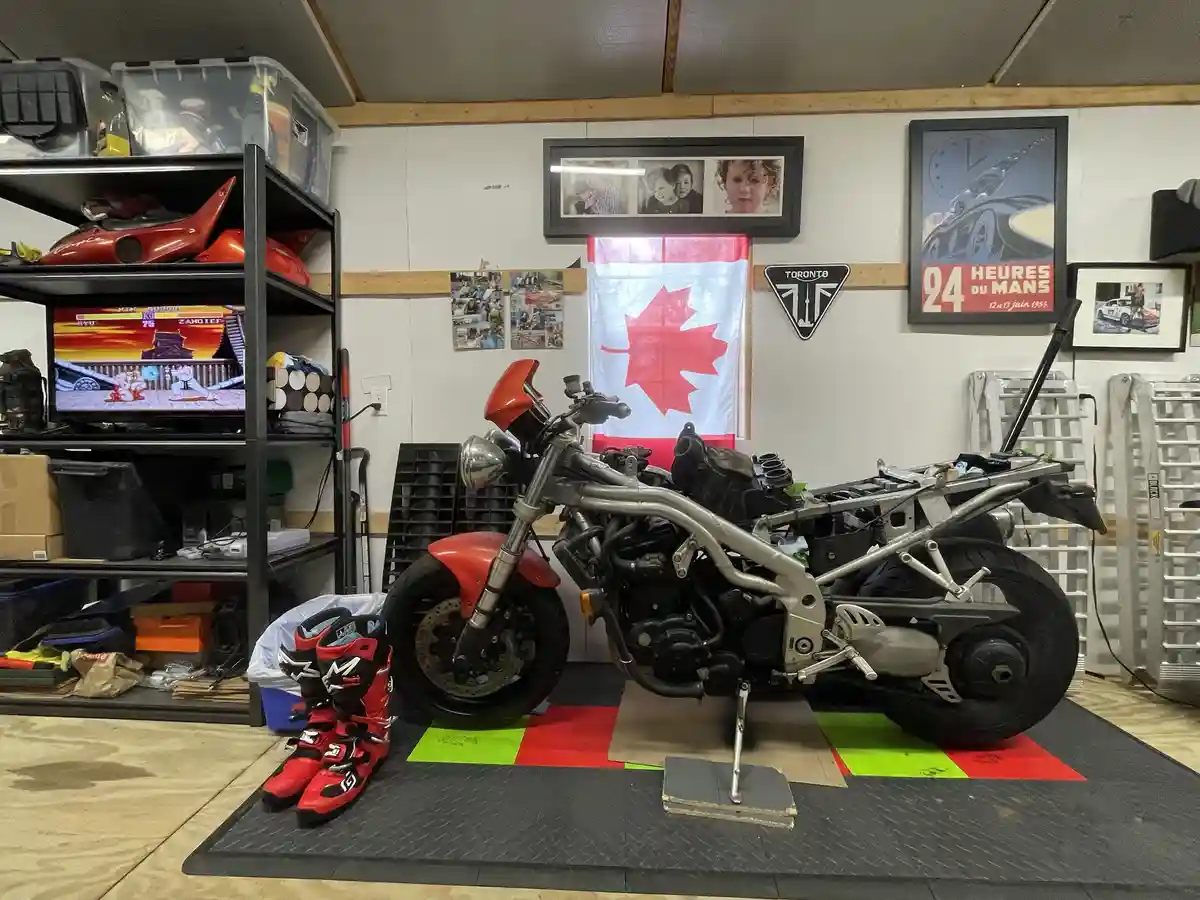 A partially disassembled red and silver motorcycle sits on a garage floor mat, surrounded by tools and gear. Behind it, a…