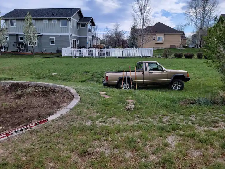 A tan pickup truck is parked on a grassy lawn in a suburban neighborhood, with gardening tools leaning against it. In the…