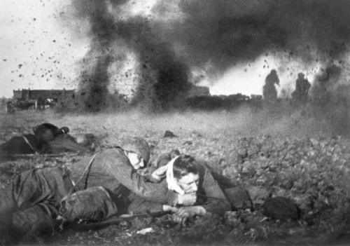 A black-and-white historical photo shows soldiers in a trench during a battle, with explosions and smoke in the background…