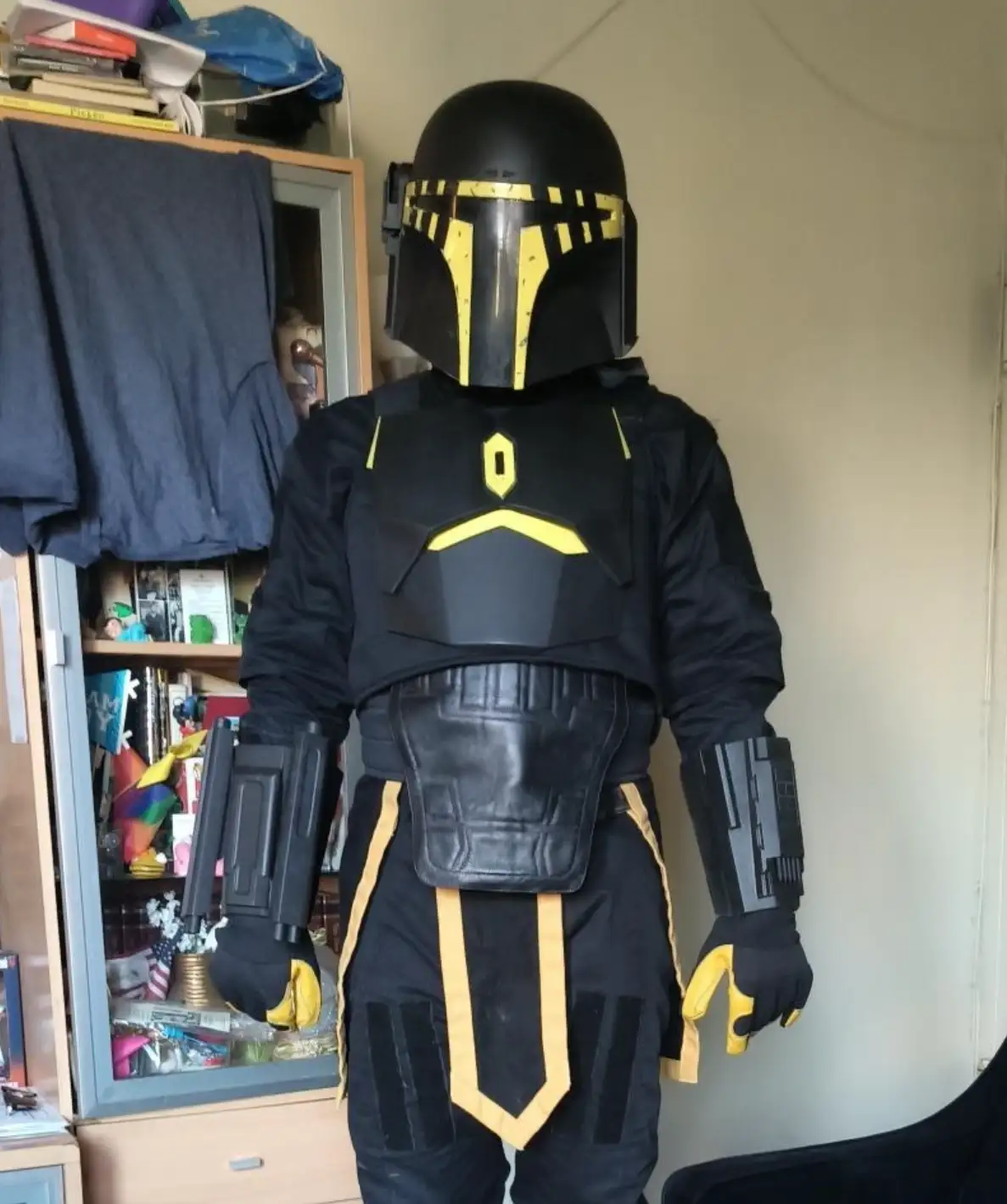 A person in a black and yellow Mandalorian-style costume stands in a cluttered room, their chest armor prominently…
