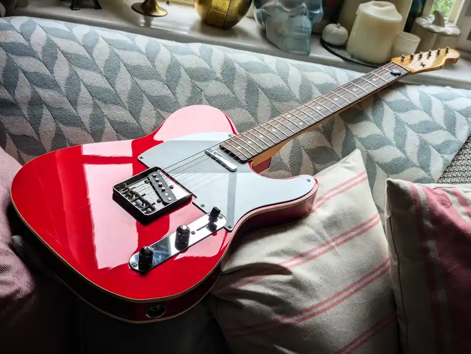 A glossy red electric guitar with a white pickguard rests diagonally across striped pillows on a patterned couch. In the…