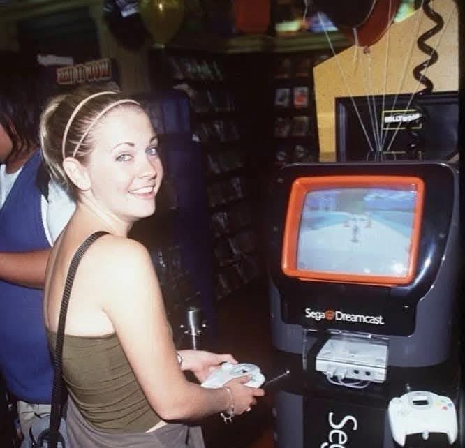 A young woman with blonde hair in a headband smiles at the camera while holding a white Dreamcast controller, standing next…
