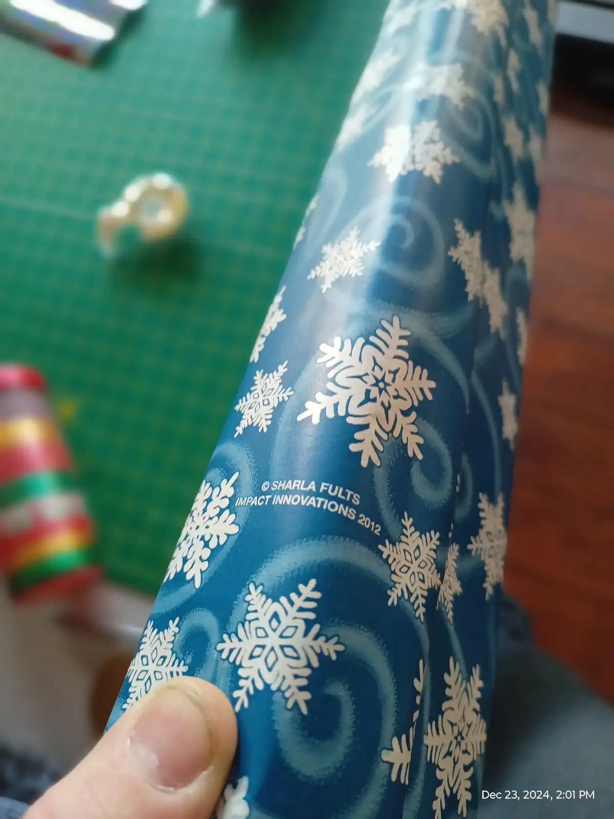 A close-up photo shows a hand holding a roll of blue Christmas wrapping paper decorated with white snowflakes and swirls…