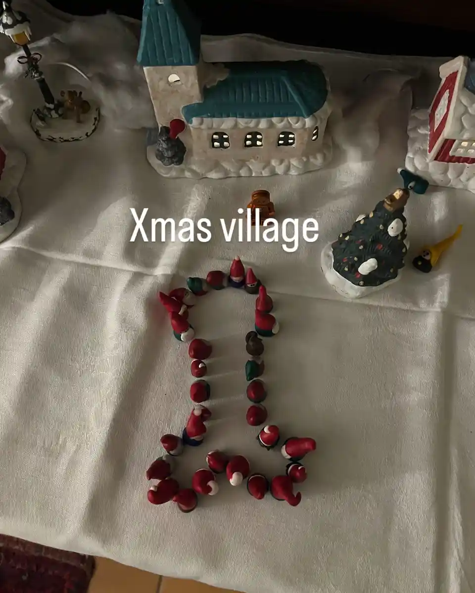 A collection of Christmas village figurines, including houses, a tree, and a lamp post, are arranged on a white cloth. In…