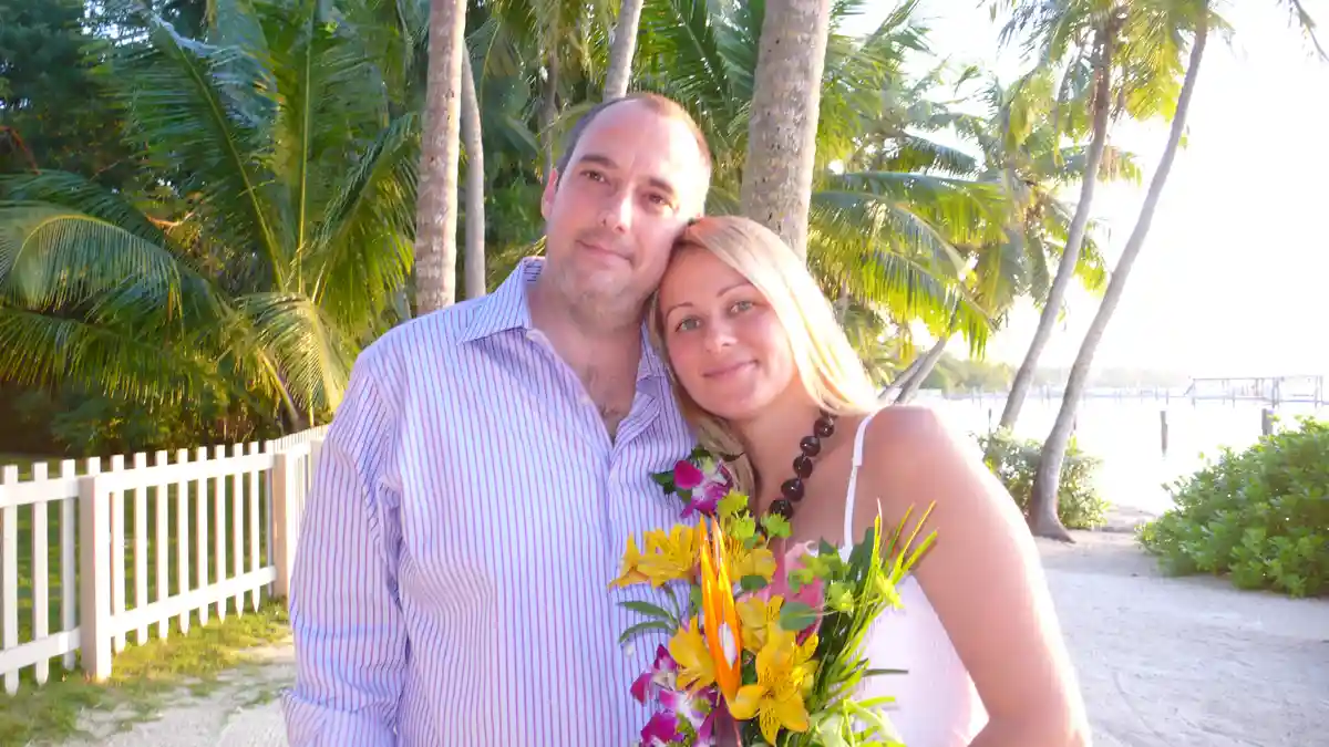 A man and woman pose together on a tropical beach path with palm trees and a white picket fence behind them. The woman…