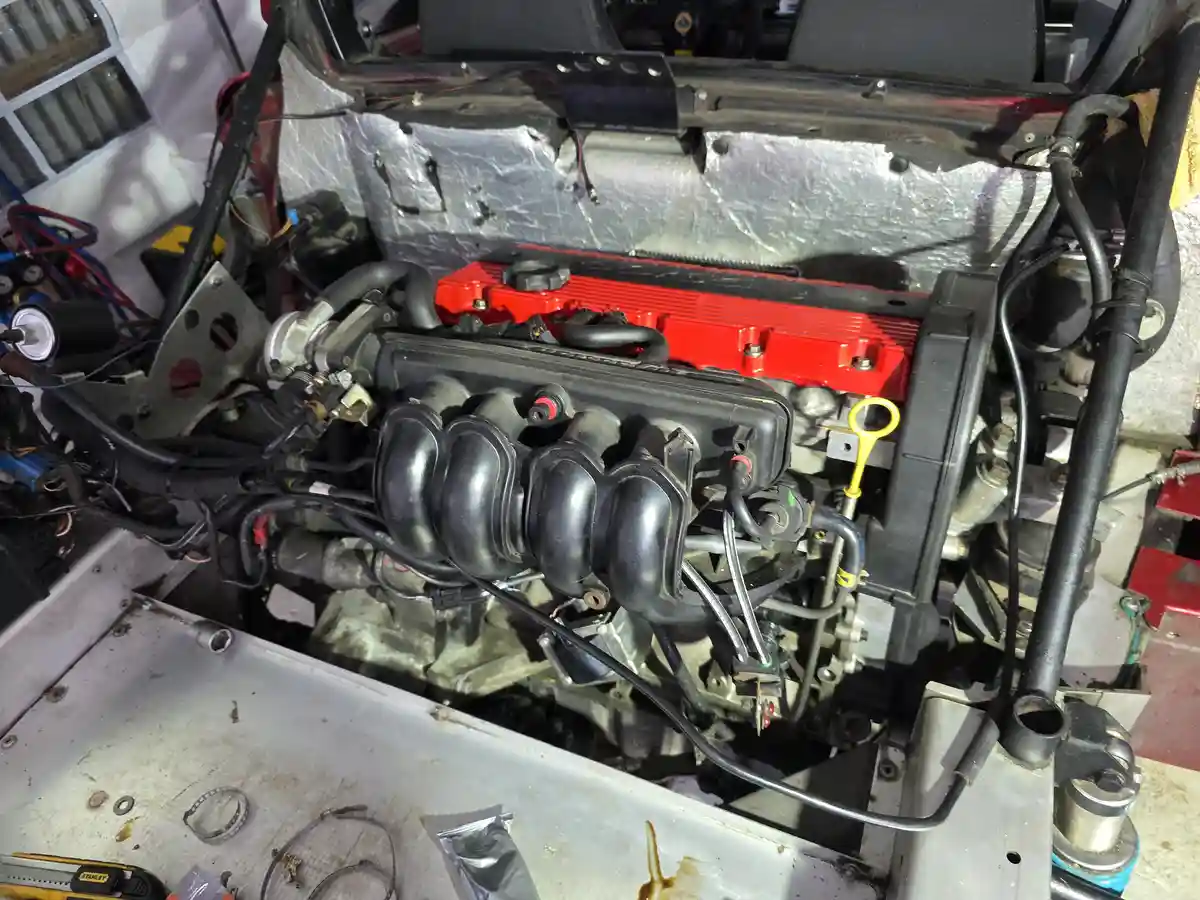 A close-up photo of a car engine bay with a red valve cover, black intake manifold, and various hoses and wires. The engine…