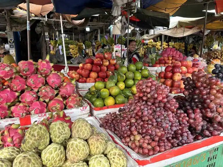 A vibrant fruit market stall displays piles of colorful produce including pink dragon fruit, green citrus, red apples, and…