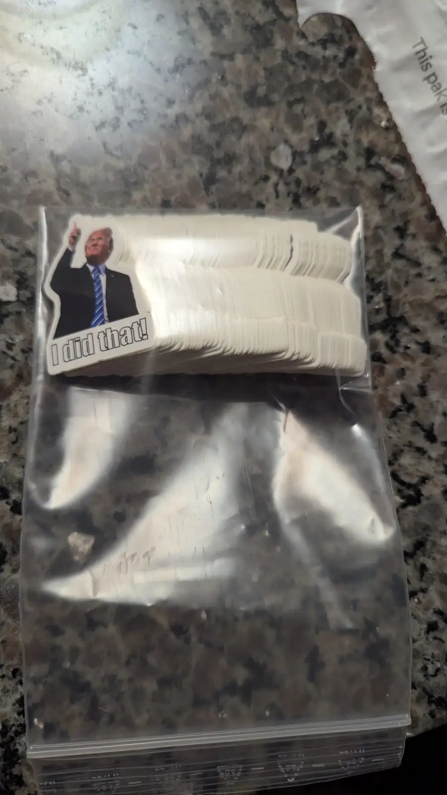 A clear plastic bag contains a large stack of identical white stickers. The top sticker features a photo of Donald Trump…