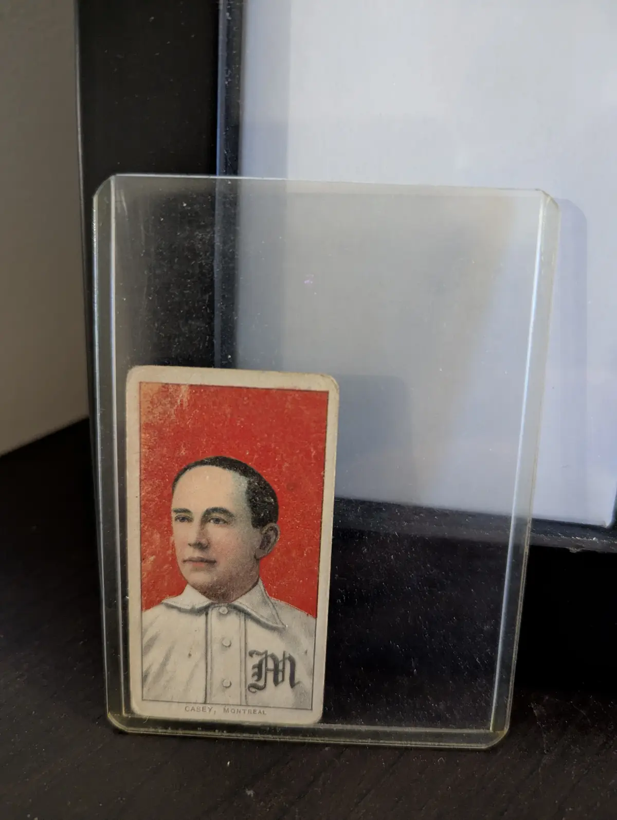 A vintage baseball card of Casey, Montreal, is displayed inside a clear plastic protective sleeve, standing upright on a…