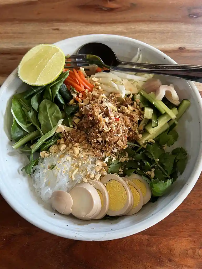 A top-down photo of a white ceramic bowl filled with a colorful Asian-style salad on a wooden table, containing sliced…