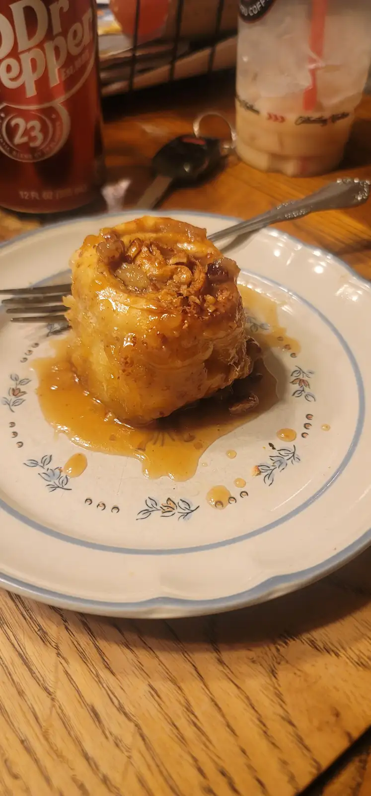 A close-up photo shows a single, generously glazed cinnamon roll with pecans on a white plate with blue floral patterns…