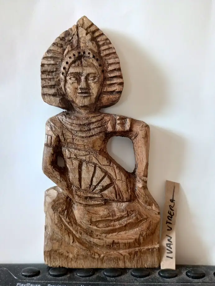 A wooden carving of a seated figure with a stylized headdress and detailed robes, mounted on a white wall next to a small…