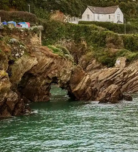 A scenic coastal photo shows a natural rock archway over turquoise water, with a white house perched on a cliff above. The…