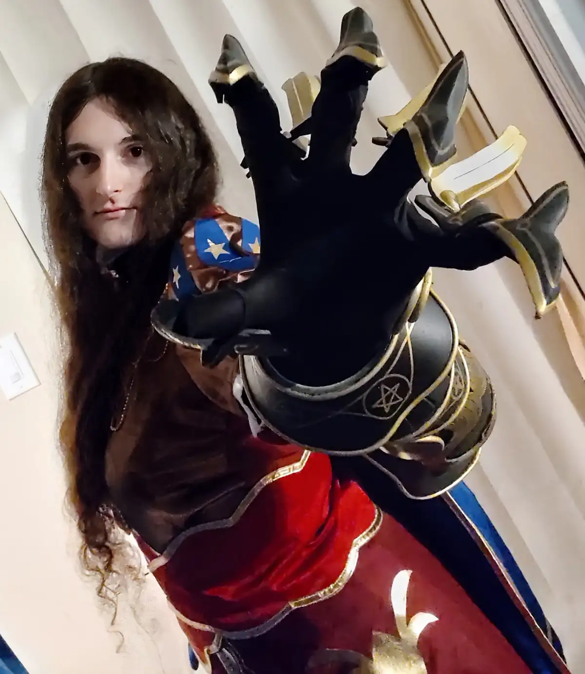 A person with long dark hair, dressed in a fantasy costume with red velvet, gold trim, and a black gauntlet featuring sharp…