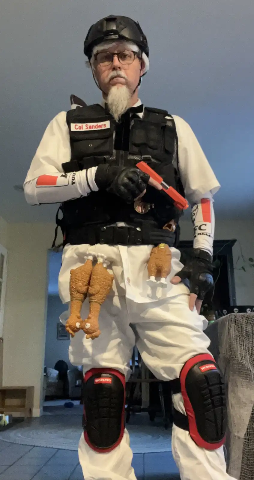 A man dressed as Colonel Sanders in a tactical outfit, complete with a helmet, vest, and knee pads, holds a toy gun while…