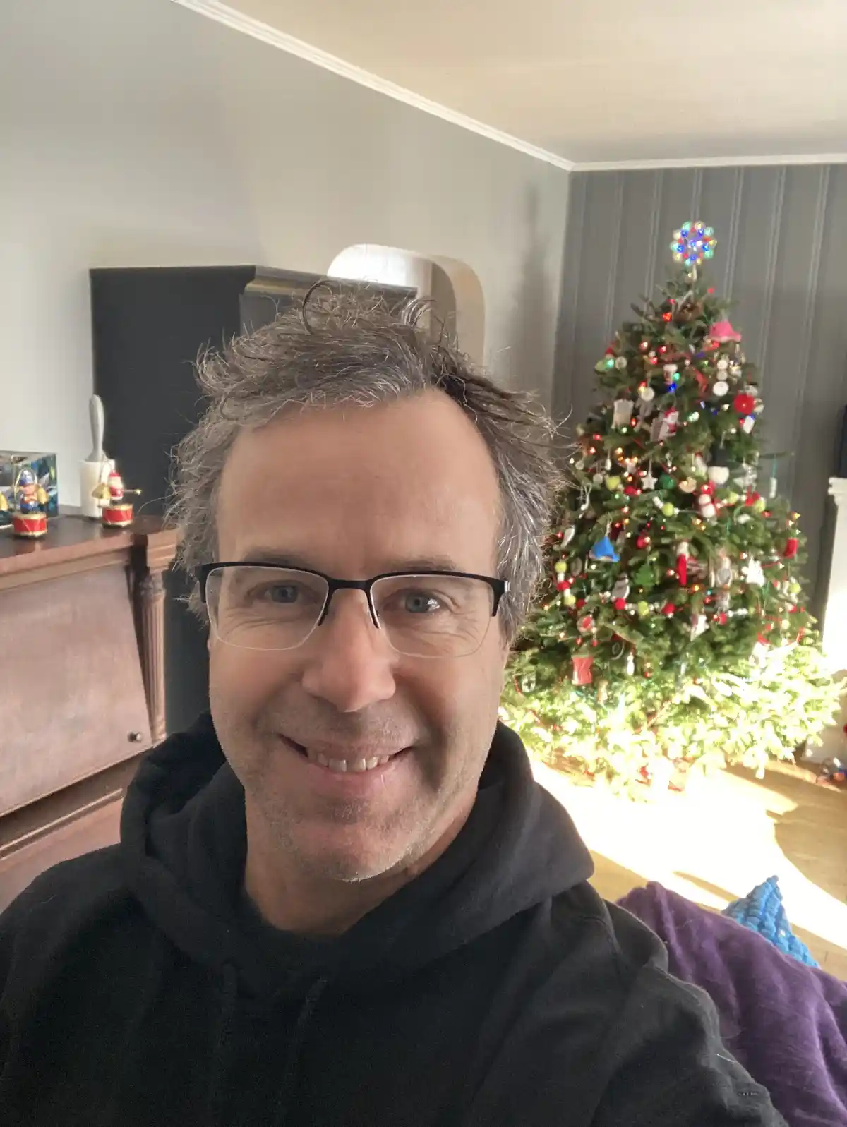 A man with glasses and a black hoodie smiles at the camera in a living room, with a brightly lit, heavily decorated…