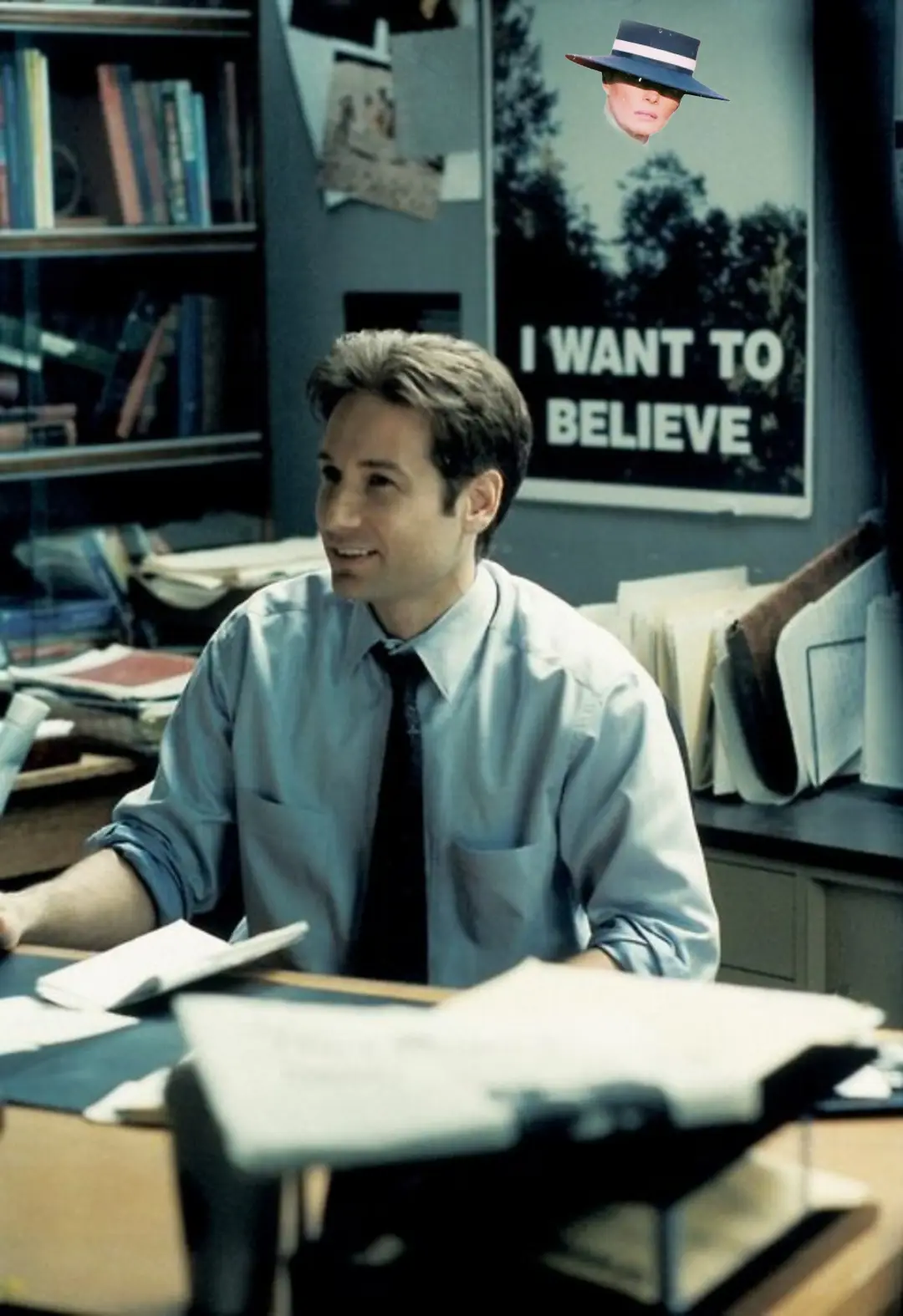 A man in a light blue shirt and tie sits at a cluttered desk in an office, smiling slightly while looking off-camera. Behind…