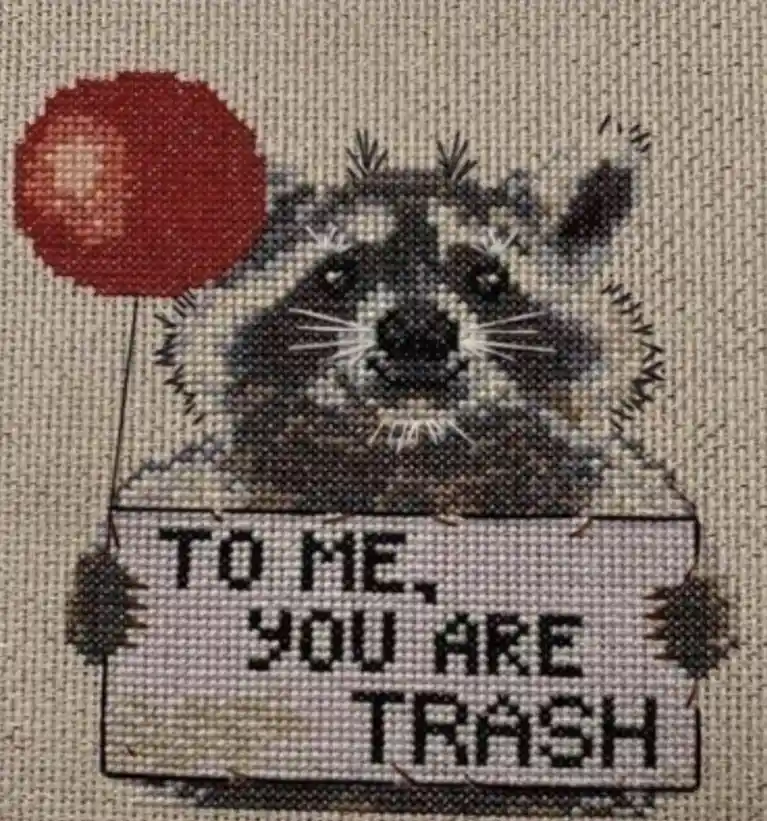A cross-stitch pattern depicts a raccoon holding a sign that reads 'TO ME, YOU ARE TRASH' while holding a red balloon. The…