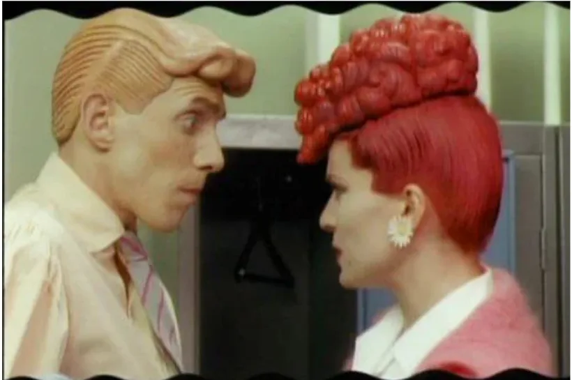 A still from a vintage TV show shows a man with a molded plastic head and a woman with a red, brain-shaped wig. They are…
