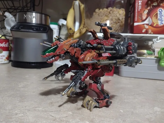 A red and orange plastic model of a dragon-like robot, heavily armed with multiple guns and cannons, stands on a kitchen…