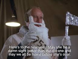 A man in a white astronaut suit with a fake white beard and mustache holds a small silver foil flag. He looks at the camera…