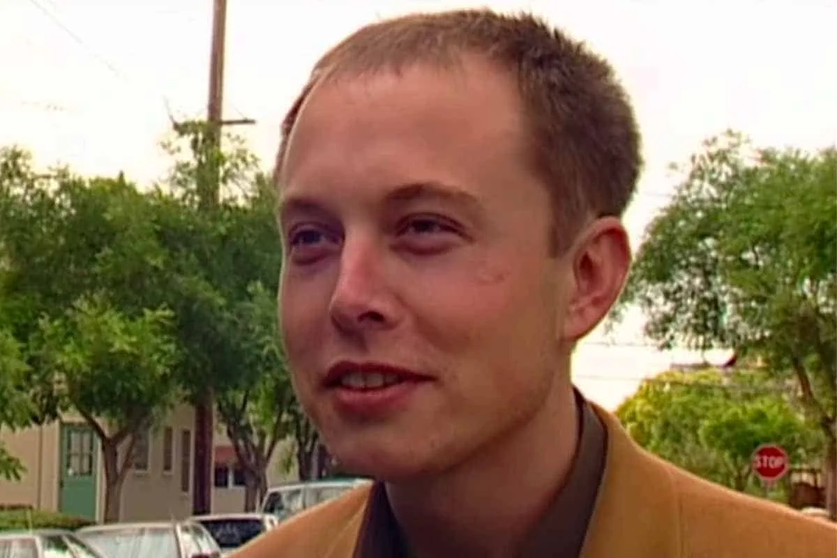 a close-up shot of a young man with short brown hair, wearing a tan jacket over a dark shirt, standing outdoors with trees…