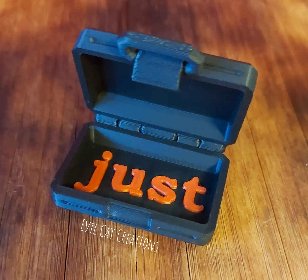 A small blue 3D-printed toolbox sits open on a wooden surface, revealing the orange 3D-printed word 'just' inside. A…