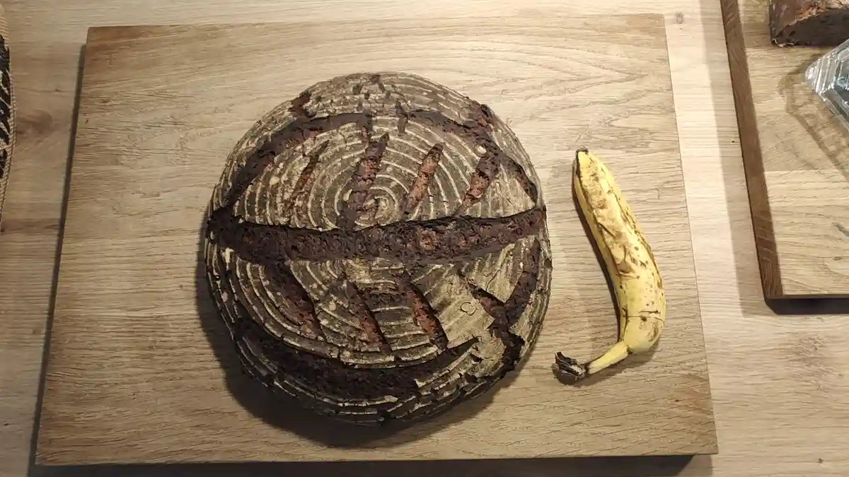 A round, dark brown loaf of bread with a cracked, swirly surface sits on a wooden cutting board next to a ripe, spotted…
