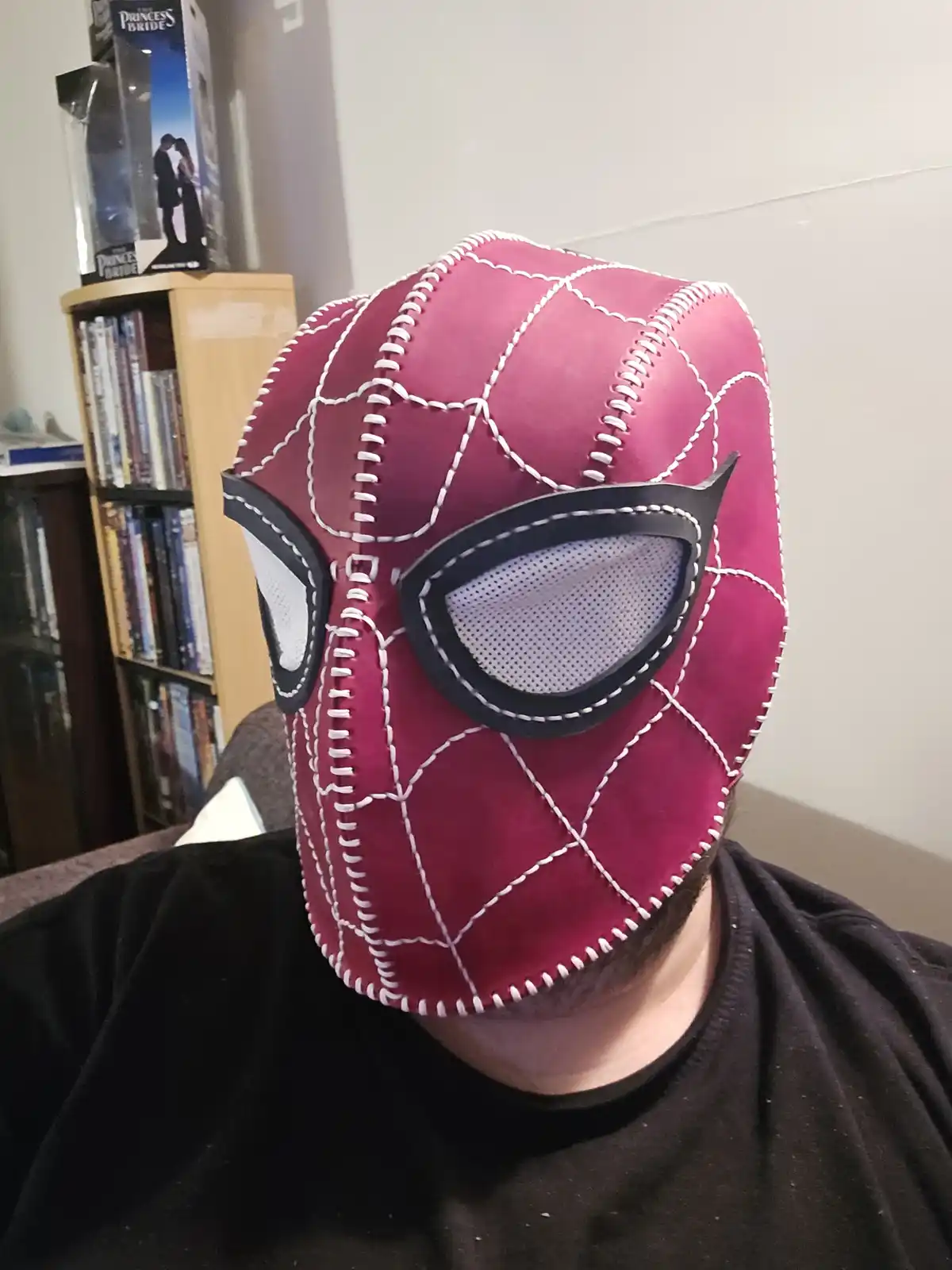 A person wearing a handmade, pink, stitched Spider-Man mask with black eye lenses and white mesh, sitting indoors with a…
