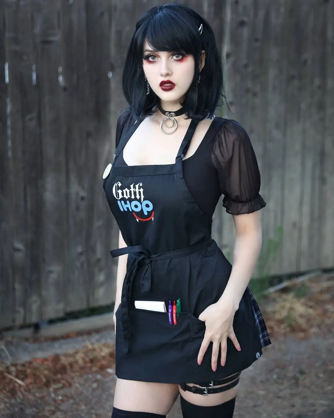 A person with black hair, gothic makeup, and a choker wears a black apron with the text 'Goth IHOP' and a vampire smile…