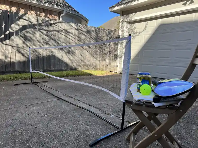A pickleball setup in a driveway with a net, paddle, and bright yellow ball resting on a folding chair next to a jar of…