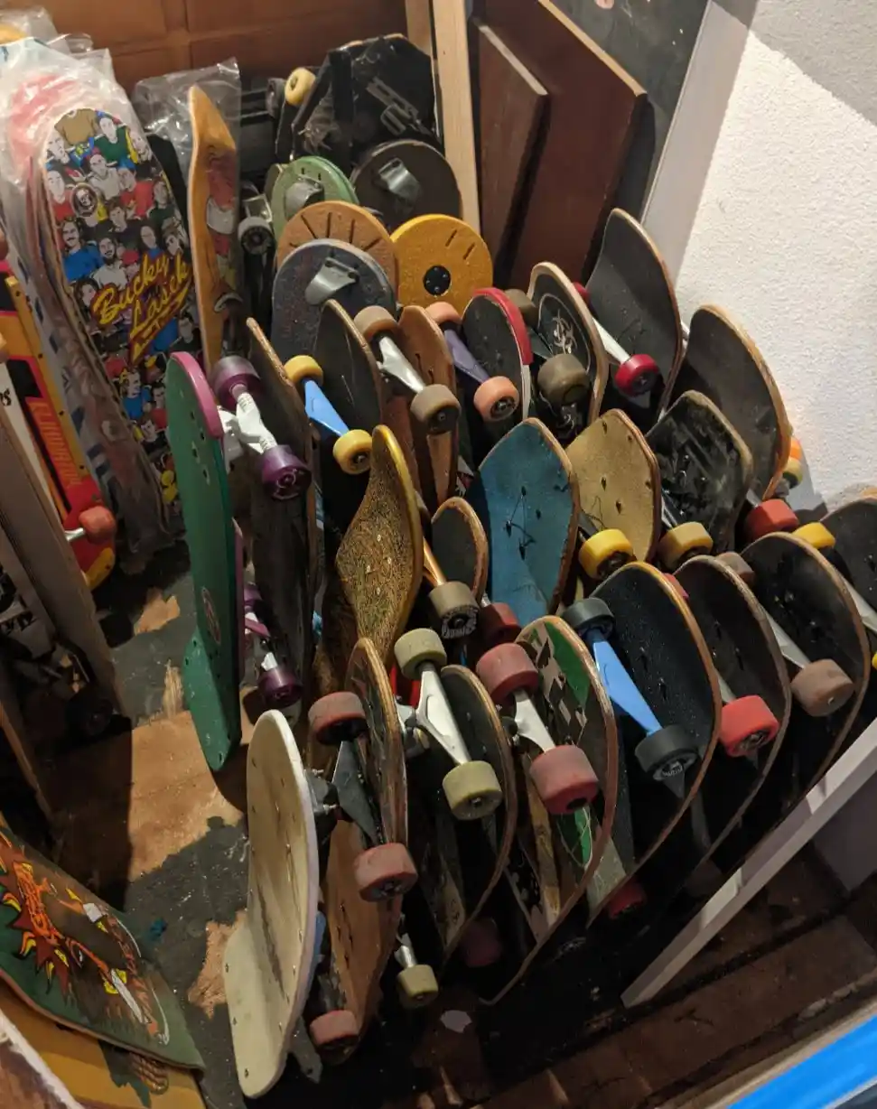 A dense, cluttered pile of dozens of skateboards, mostly showing their undersides with worn wheels and trucks, leaning…