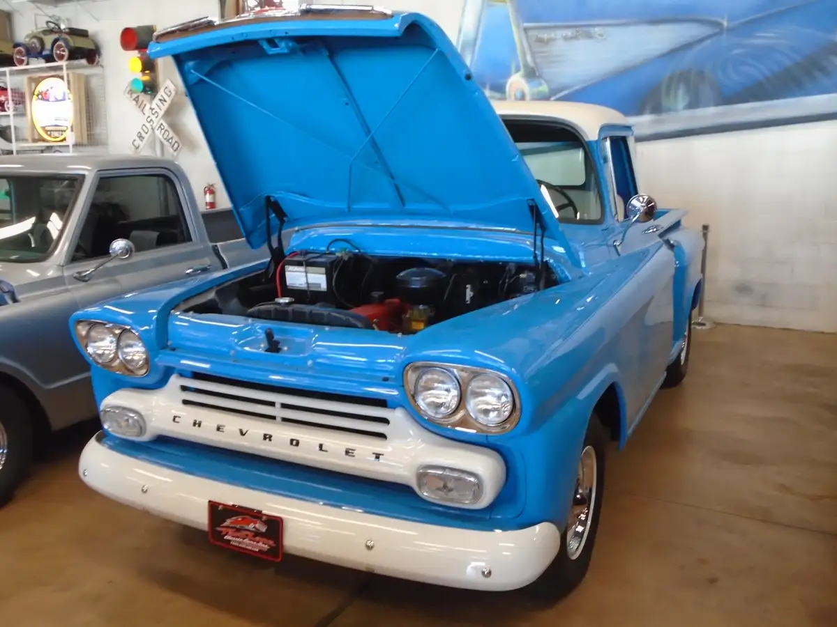 A vintage blue and white Chevrolet pickup truck with its hood open, revealing the engine, is parked indoors in a garage or…