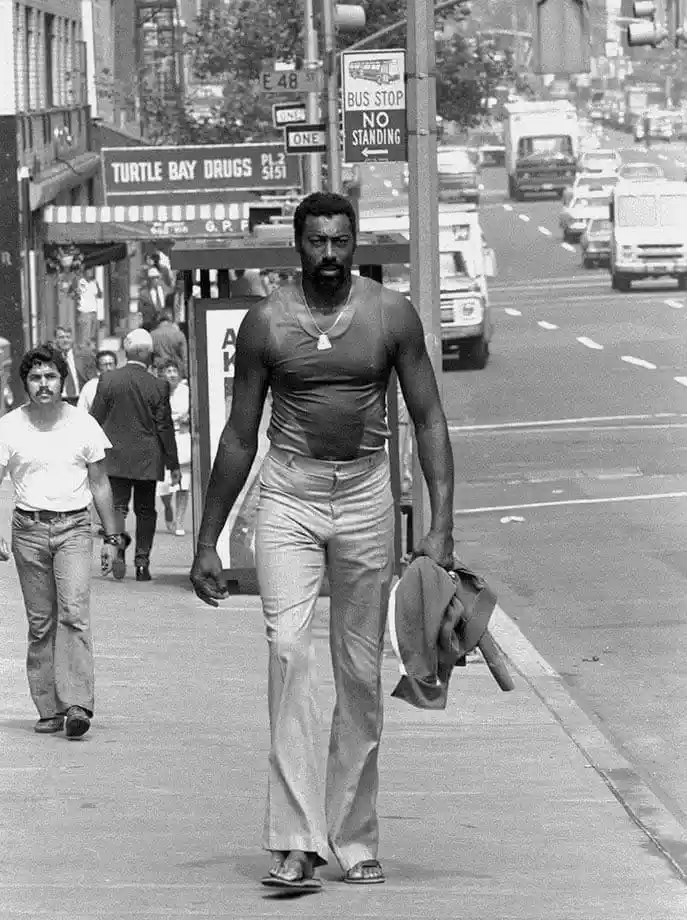 A black-and-white photo shows a muscular Black man walking down a city sidewalk, wearing a tight tank top, wide-leg pants…