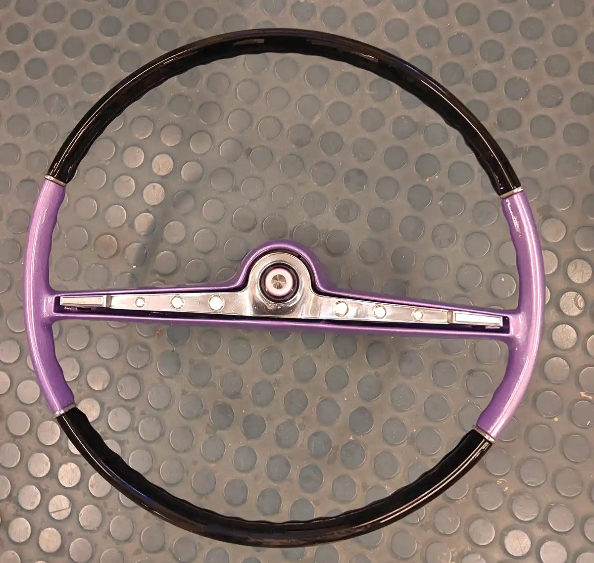 A vintage-style car steering wheel with a glossy purple center and black outer rim rests on a gray, dimpled metal surface…