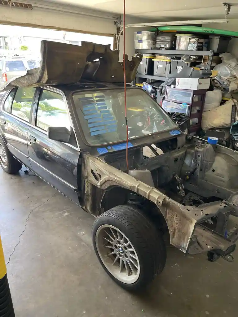 A black BMW sedan is shown in a garage with its front end stripped down to bare metal, the hood removed and propped up, and…