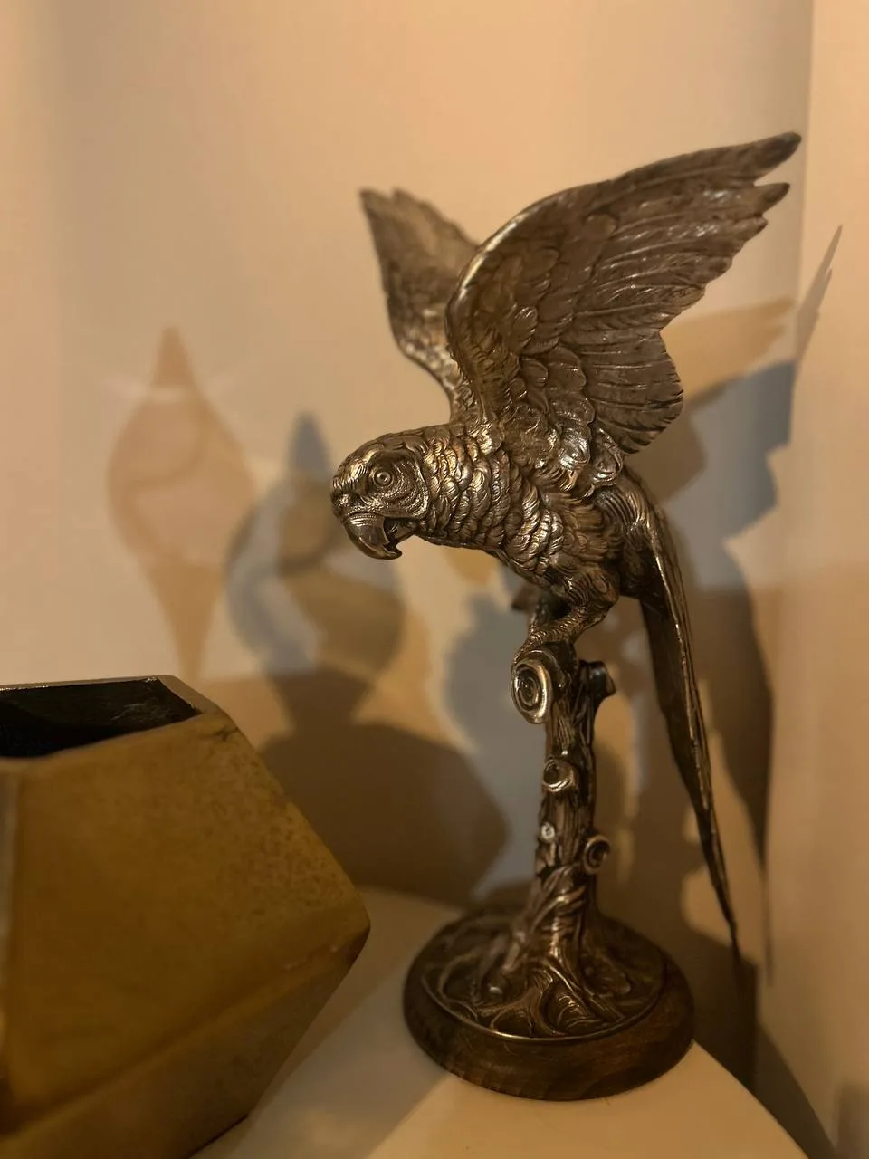A detailed silver-toned metal statue of a parrot with wings spread, perched on an ornate branch-like stand, positioned on a…