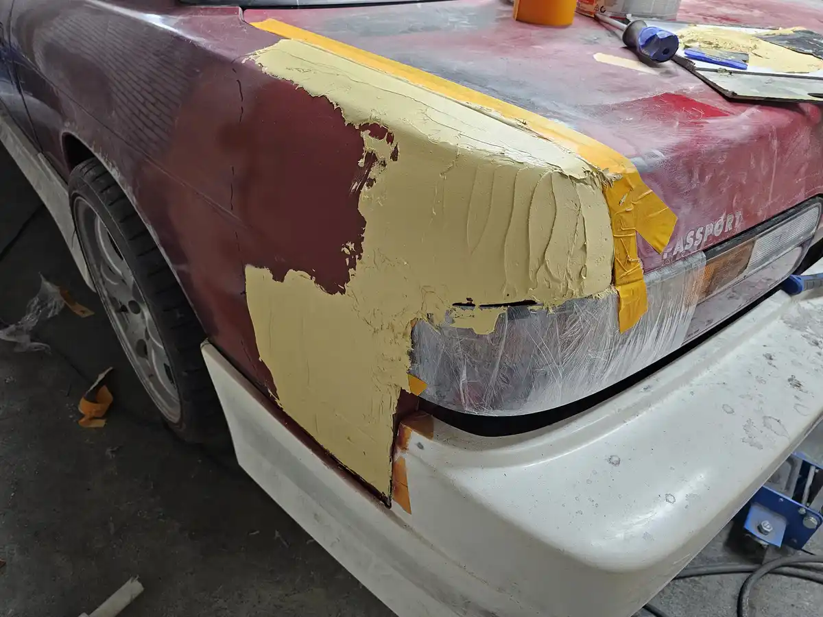 A close-up of a maroon car's front fender and headlight area, where thick, uneven beige paint has been applied over a…