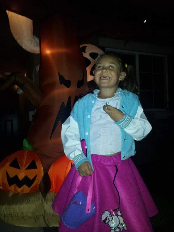 A young girl in a light blue jacket and purple skirt stands in front of Halloween decorations, including a large inflatable…