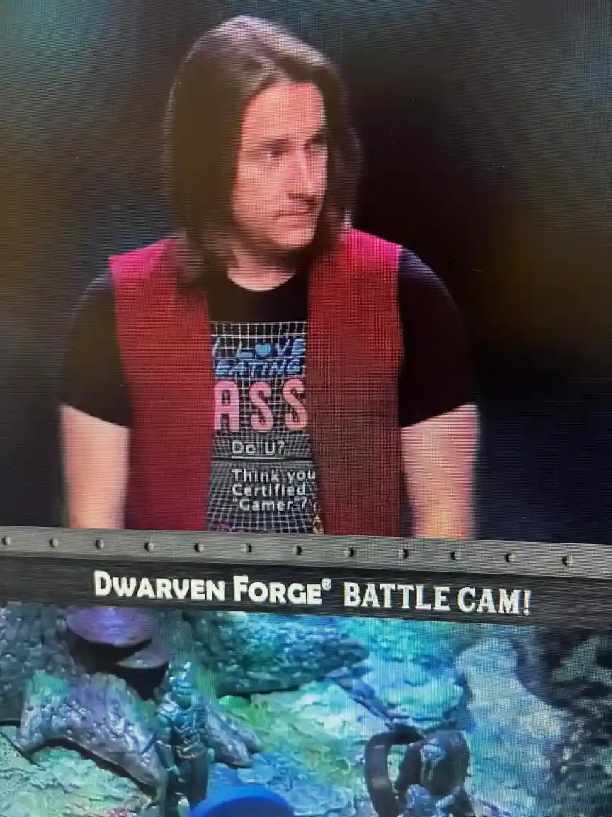 A man with long brown hair wearing a red vest over a black t-shirt that says 'I LOVE EATING ASS DO U? Think you Certified…