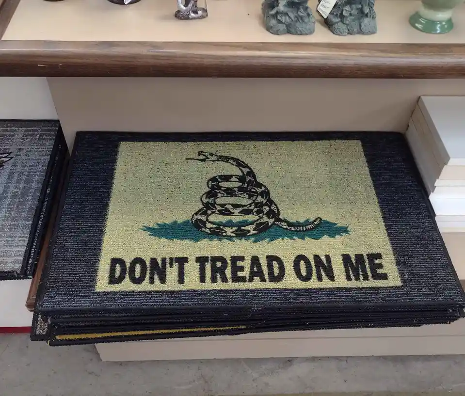 A folded doormat featuring the Gadsden flag design with a coiled rattlesnake and the text 'DON'T TREAD ON ME' is displayed…