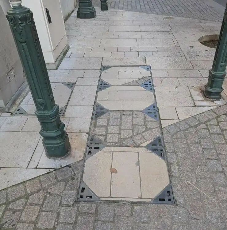 A photo of a sidewalk with a rectangular metal grate embedded in it, surrounded by light-colored stone tiles and darker…