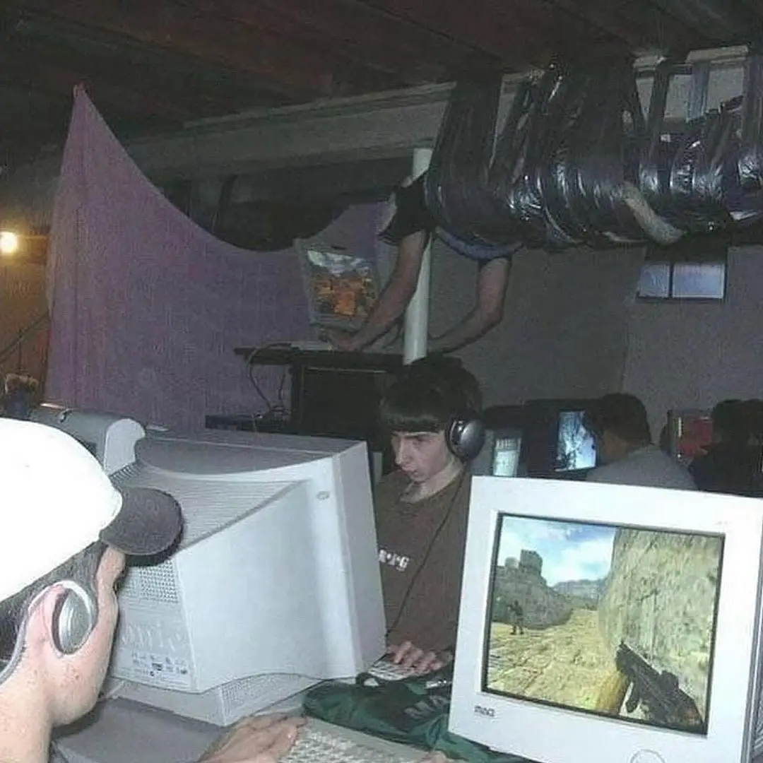 A dimly lit room with multiple people playing old-style computer games on bulky CRT monitors. One person in the foreground…