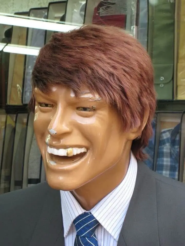 A mannequin dressed in a suit and tie with a wide, toothy grin has a cigarette butt stuck between its teeth and a small…