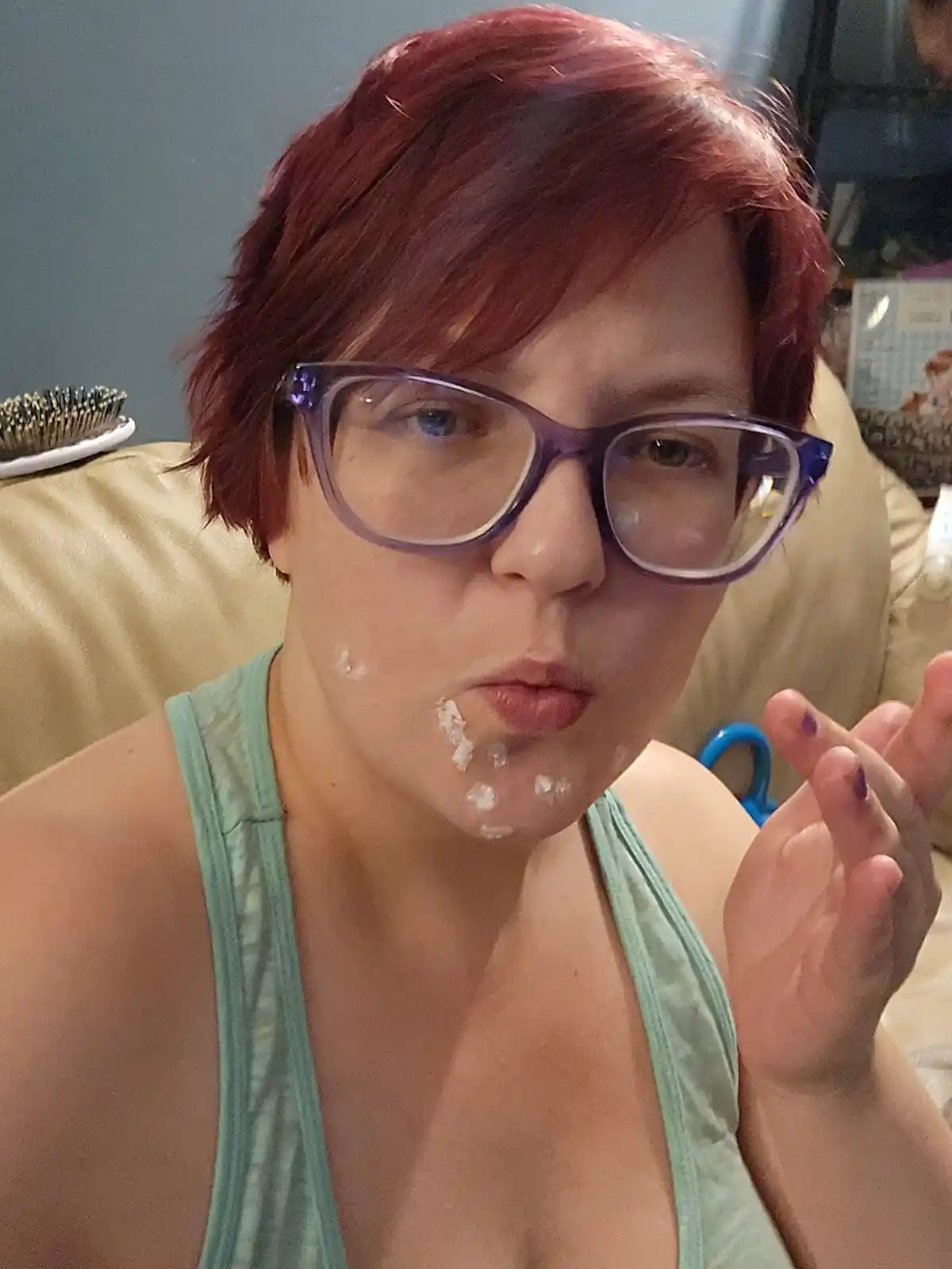 A person with short red hair and purple glasses sits on a beige couch, making a pouty face while white shaving cream is…