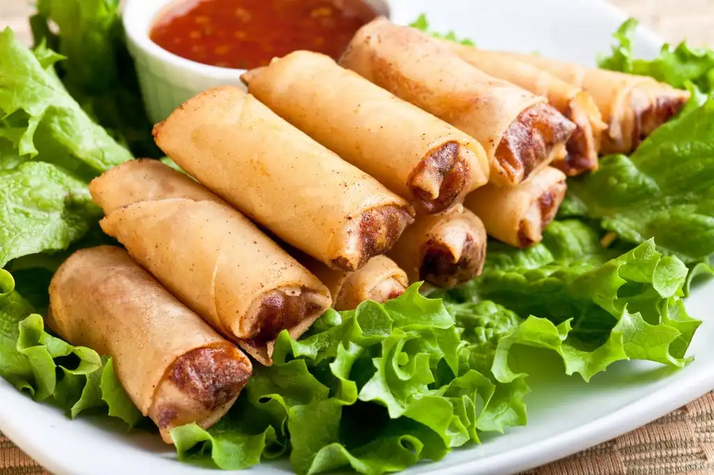 A plate of golden-brown fried spring rolls arranged on a bed of fresh green lettuce, with a small bowl of red dipping sauce…