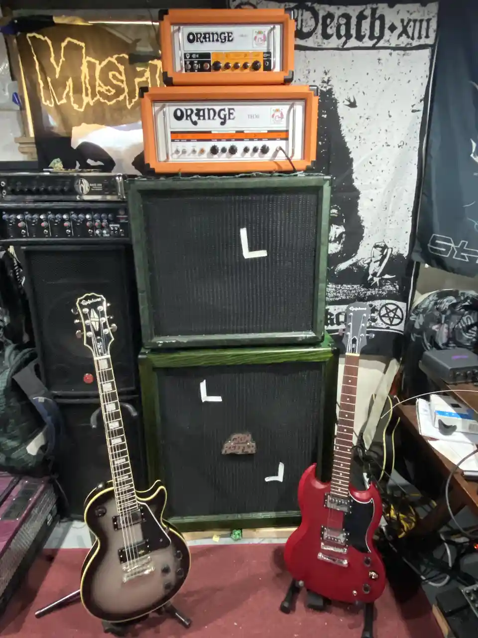 Two electric guitars stand on stands in front of a stack of amplifiers, including two orange head units and two green…
